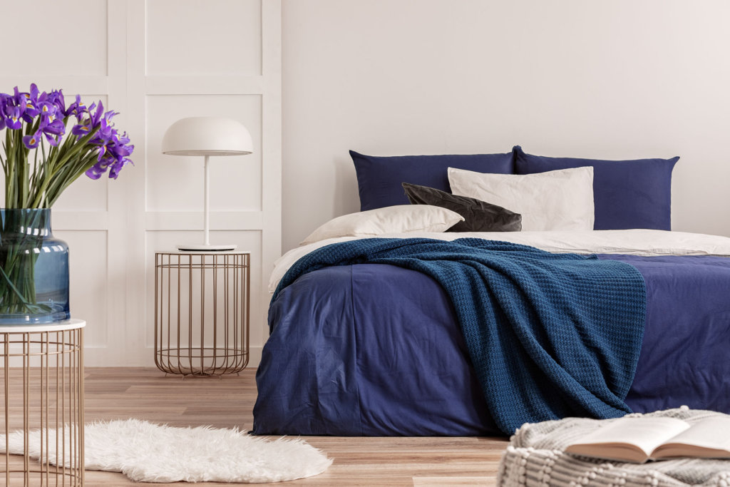 Purple flowers in blue glass vase on stylish table in white bedroom interior with comfortable bed