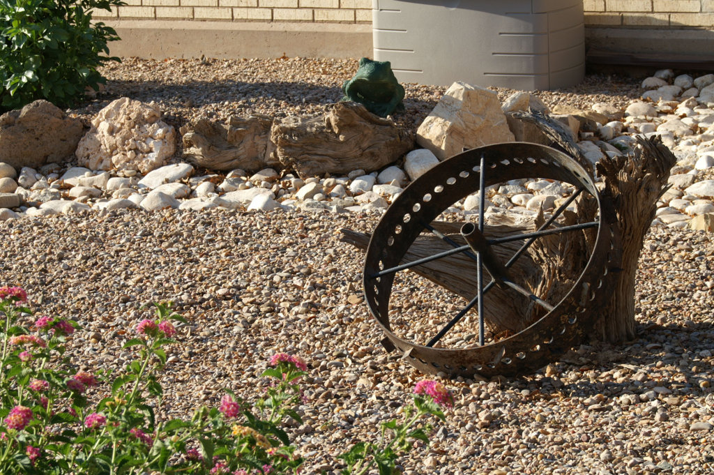 Old metal wagon wheel lying amidst pebbles, driftwood, and other yard decorations as part of a desert landscaping theme.