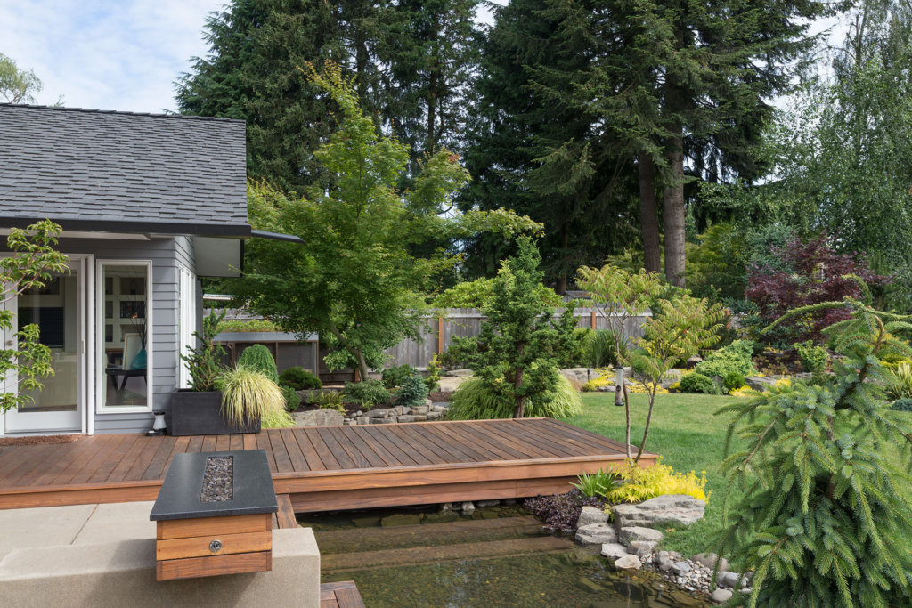 Back yard of a contemporary Pacific Northwest home featuring a deck spanning a creek-like water feature with a landscaped lawn in the background.